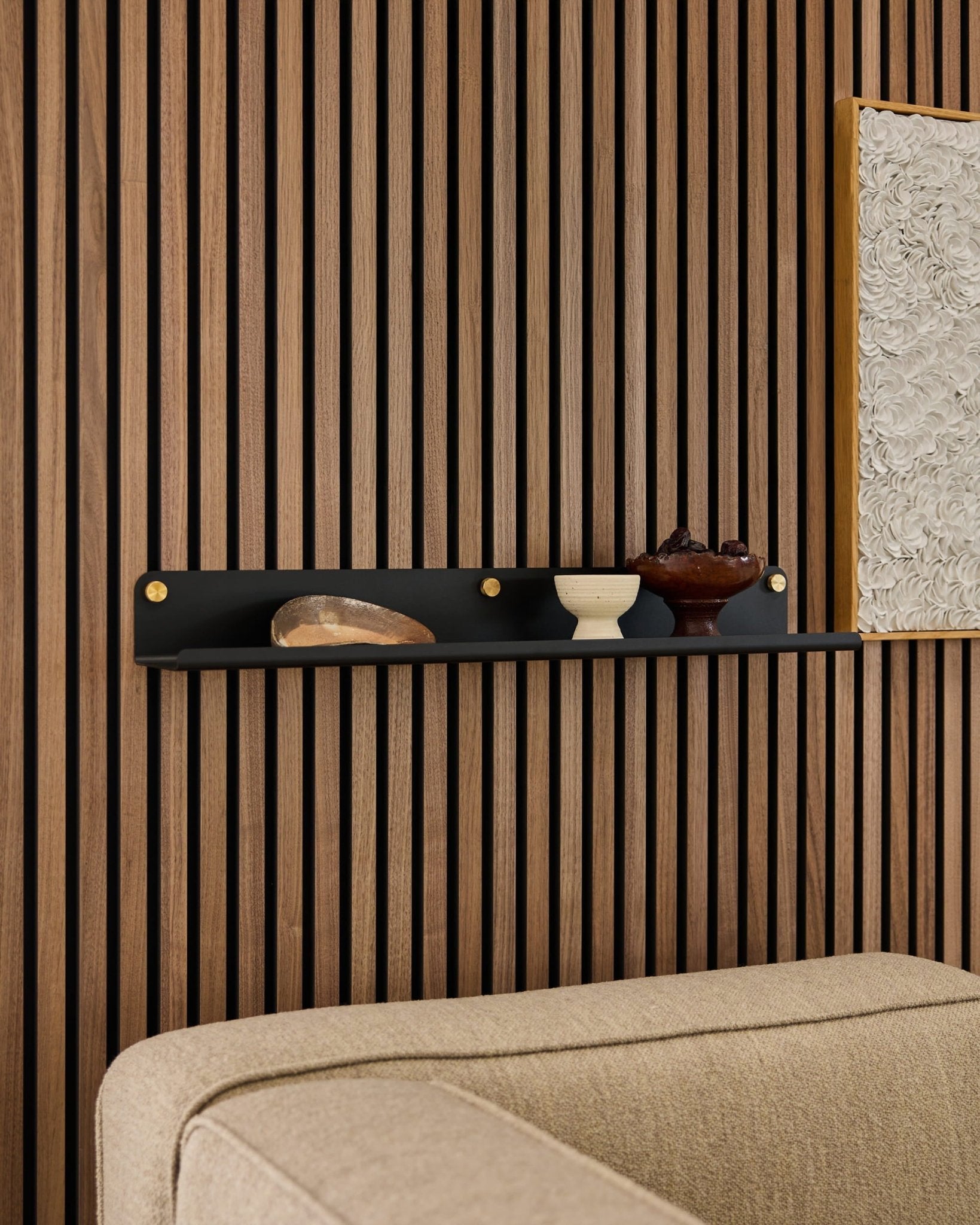 Étagère large WoodUpp Norr noire (75,9 cm) agrémentée déléments décoratifs, placée au-dessus dun canapé beige, fixée sur un panneau mural moderne en bois à lattes acoustiques.